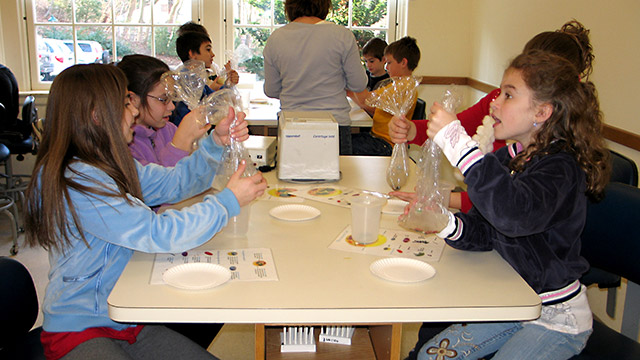 Phtotgraph of several elementary-aged students at lab tables holding clear plastic bags that contain materials that represent parts of a human cell.