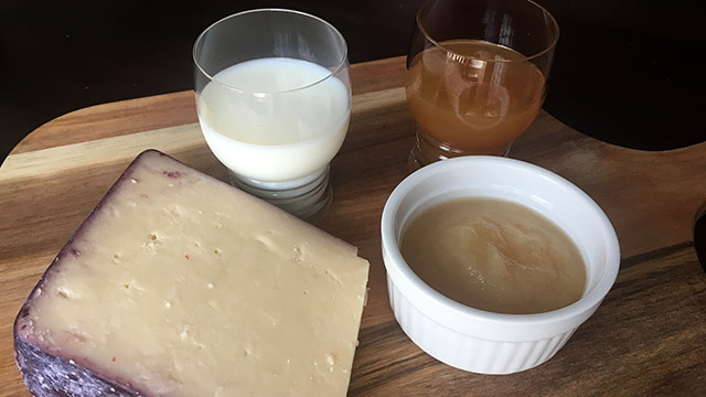 Wood cutting board with glasses of milk and apple juice, a bowl of apple sauce, and a block of light-colored cheese.