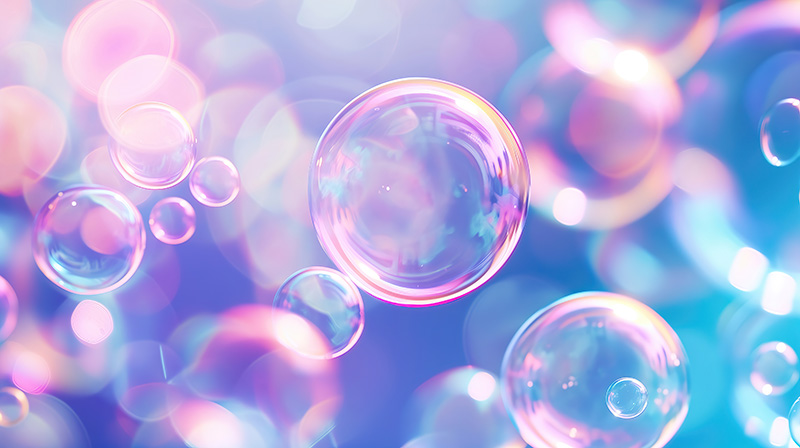 A soft focus background of bokeh lights in shades of pink, purple, and blue frames several translucent soap bubbles, catching light and showing iridescence. AdobeStock_721355718