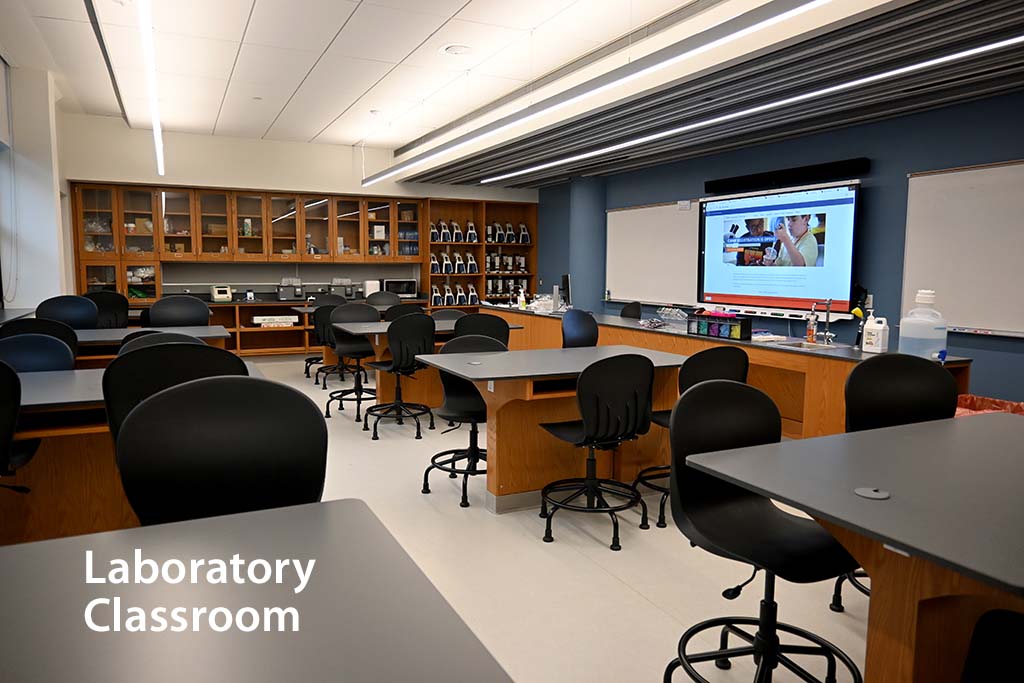 Laboratory classroom with eight desks and an intstructor counter in front of a white board and smartboard monitor. Shelves in the background hold microscopes and other scientific supplies