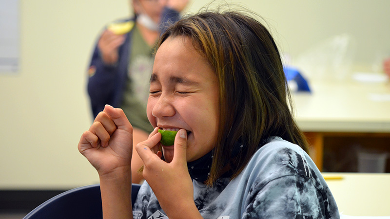 A girl with dark hair and a tie-dye shirt makes a funny face as she bites into a small green lime. Her eyes are squeezed shut and her mouth is open wide.