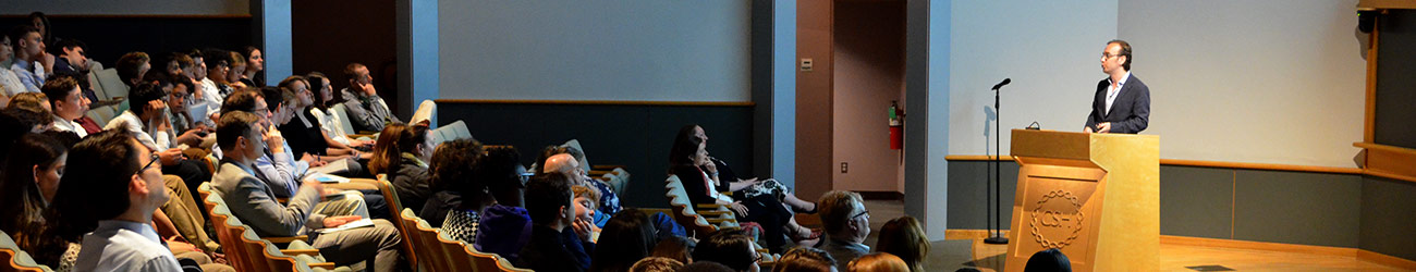 Scientist at poium in an auditorium with several seated people looking on