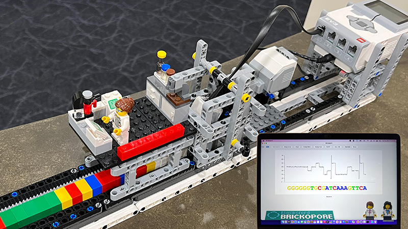 A LEGO sequencer, designed to resemble a scientific instrument, is displayed on a table. In the foreground, a laptop screen displays scientific data, including a graph and a sequence of genetic code.