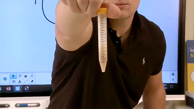 Out of picture frame person wearing a black shirt holds a test tube up to the viewer containing a pale yellow substance.