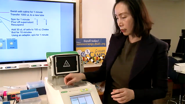Person with shoulder length dark hair wearing dark clothing places a test tube into a rectangular PCR machine. Digital board in background displays text.