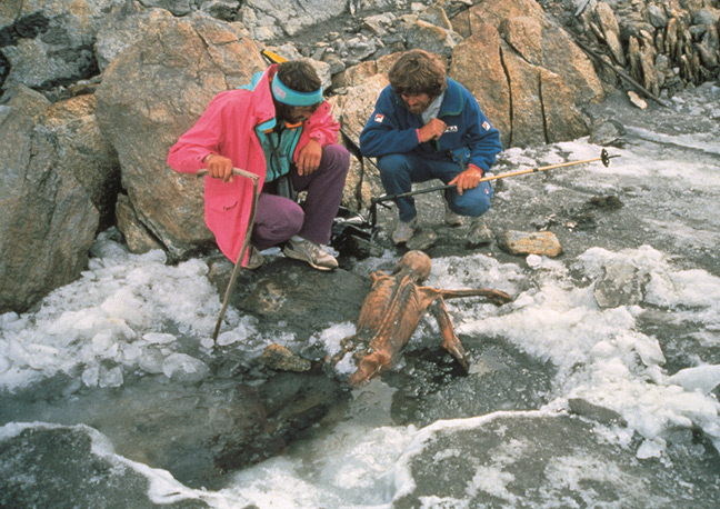 two people looking at Otzi mummy embedded in slushy icy  ground in mountainous setting. Copyright Paul Hanny