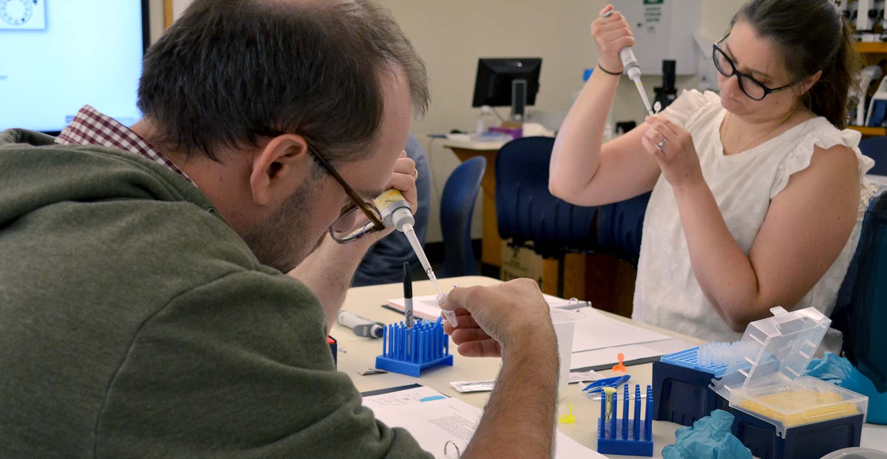 Two adult person each holds a microtube and a pipette at a desk with scientific supplies in a laboratory classroom.